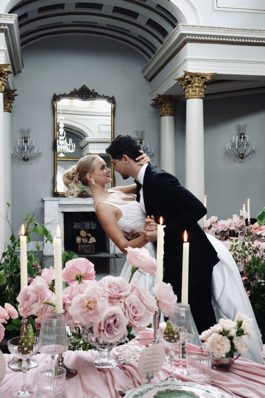 Bride and groom beside a wedding table styled with pink flowers, glassware and white taper candles
