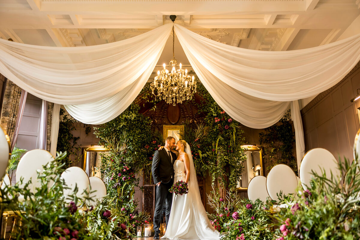 Bride and groom beneath ivory ceiling drapes, surrounded by abundant flowers and foliage